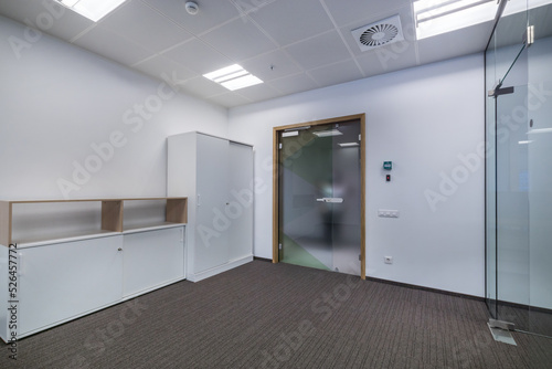 A bright  empty office space with gray flooring  white cabinets and a glass door.