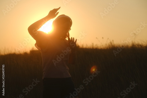 Beautiful woman in headphones and glasses dancing at sunset in the countryside at sunset in casual clothes