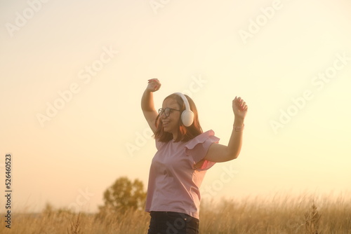 Beautiful woman in headphones and glasses dancing at sunset in the countryside at sunset in casual clothes