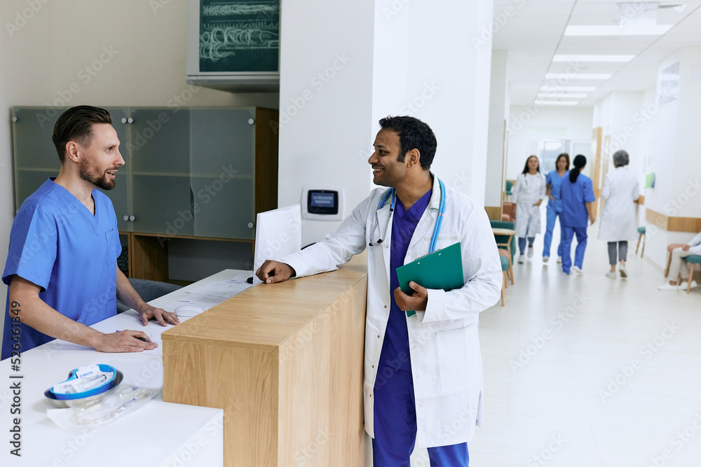 Fototapeta premium Indian doctor talking to male nurse near reception desk at modern hospital while working day. View of lobby medical clinic with patients and medical personnel