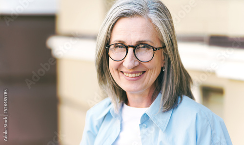 Charming grey hair mature woman wearing eye glasses standing outdoors enjoying vacation traveling around world at retirement. Mature woman with perfect skin wearing blue shirt