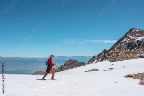 Tourist in red walks on snow mountain near abyss edge on high altitude under blue sky in sunny day. Man with camera on snowy mountain near precipice edge with view to large mountain range in away. © Daniil