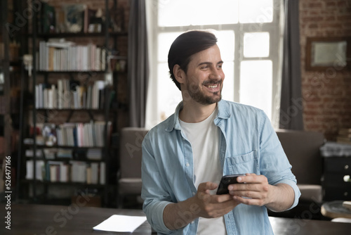 Cheerful dreamy millennial smartphone user man holding cell, standing at work table in home office, looking away with happy smile, feeling inspired, thinking, enjoying wireless online communication