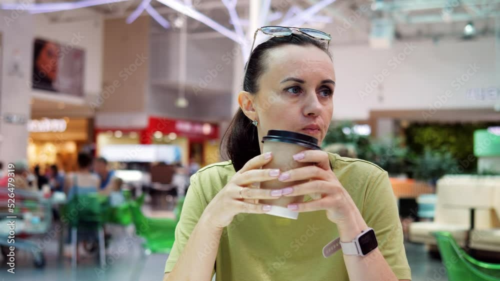 A woman at a food court table in a shopping mall drinks coffee from a cardboard cup. A woman at a food court table in a shopping mall drinks coffee from a cardboard cup.