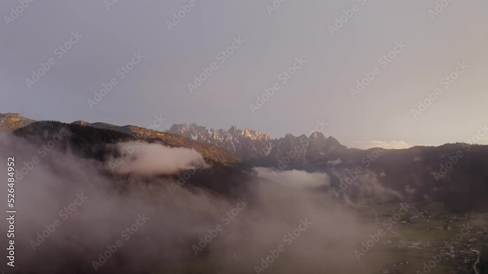 Panoramic view of a mountain valley with a village lying in a lowland. The light of the rising sun illuminates the peaks of the rocky mountains. Austria, Alps, drone video through the clouds Panoramic view of a mountain valley with a village lying in a lowland. The light of the rising sun illuminates the peaks of the rocky mountains. Austria, Alps, drone video through the clouds