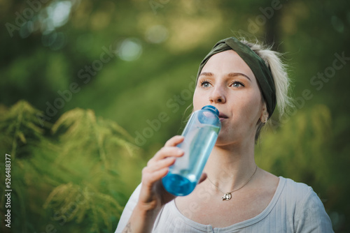 closeup o f a young healthy alternative & spiritual looking woman drinking fresh detox water from a blue plastic bottle outside in the park with a green refreshed nature forest background