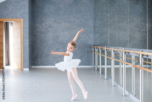 Little ballerina dancing in ballet studio. Cute child girl in white ballet shoes, dress, tutu skirt in class room. Kid in classical dance school for children. Small dancer doing exercises by barre.