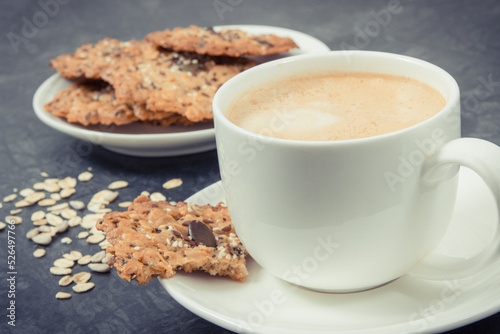 Cup of white coffee and fresh baked oatmeal cookies with honey and healthy seeds on plate. Delicious crunchy dessert