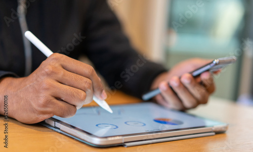 Close up focus on employee hand use pen point statistic report to review on tablet while check smart phone in office room for doing many job task concept for business technology