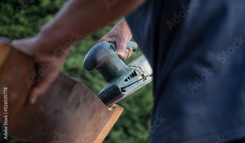 Close-up of an amateur craftsman with an orbital sander in his hand touching up a wooden board in the garden