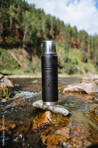 A black metal thermos stands on a stone in a mountain river, a vacuum bottle for hot drinks, tea on a hike in the mountains, a thermo flask.