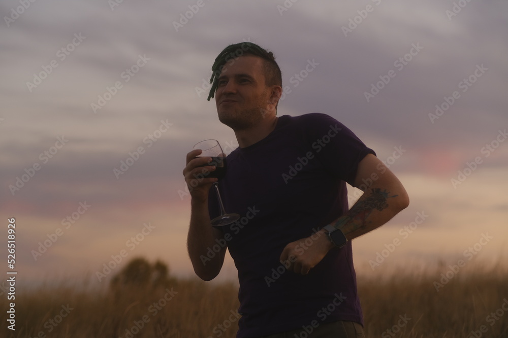 Fototapeta premium Young man dancing with glass of wine in countryside. Hipster with green dreadlocks in casual clothes resting with alcohol in nature at sunset