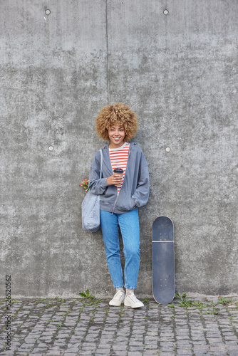 Full length shot of beautiful curly haired woman drinks takeaway coffee wears sweatshirt jeans and sneakers carries net bag poses against grey concrete wall near skateboard. Youth and lifestyle © wayhome.studio 