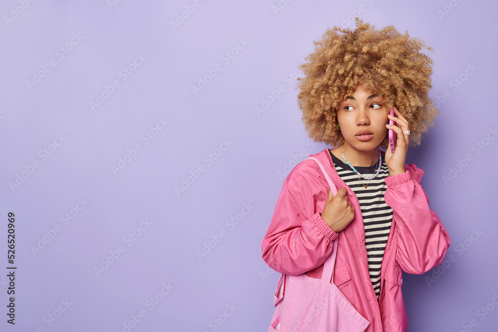 Horizontal shot of thoughtful curly haired young woman makes phone call keeps smartphone near ear discusses something concentrated away wears striped jumper and windbreaker carries fabric bag