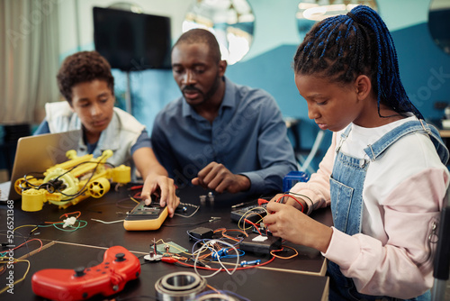 Side view portrait of young black girl building robots with male teacher in background during engineering class at school