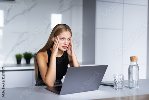 Sad woman sitting in the kitchen and working on laptop and counting spendings and taxes