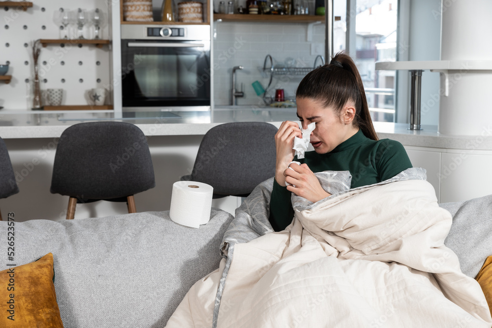 Young sick stressed woman lying at home covered with blanket blowing