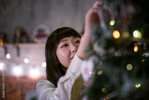 a pretty dark-haired Korean girl in a cozy Christmas room with festive bokeh lights in the evening dresses up a Christmas tree with eco-natural toys. Portrait.