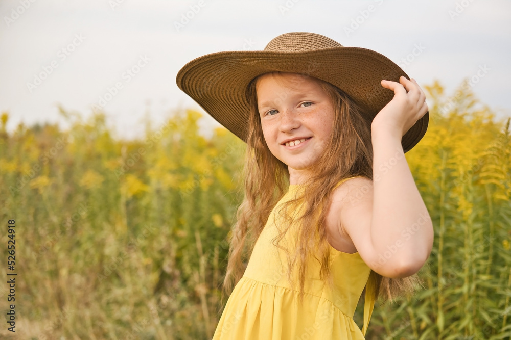 Happy and smiling cute little girl in yellow dress and hat walks in a field. Summer outdoor lifestyle. Portrait of the beautiful girl with long hair.