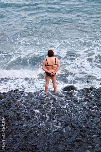 Vertical shot of a woman in bikini standing on the beach with her feet in water photo