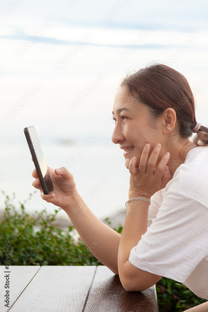 Asian women take photos and video calls at sea and beach in the morning at sunrise with their mobile phones, smartphones for posting to the internet online while traveling on vacation.