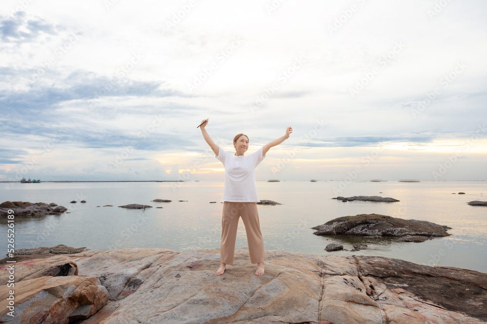 Asia woman with smart phones relaxing and arm up on beach. woman stretch relax breathe fresh air. Happy traveler asian woman relieve negative emotions enjoying lazy morning, having peaceful weekend.