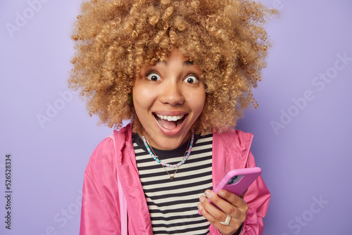 Modern technologies and mobile apps concept. Positive surprised woman with curly hair holds mobile phone feels happy chats online wears striped jumper and pink jacket isolated over purple background