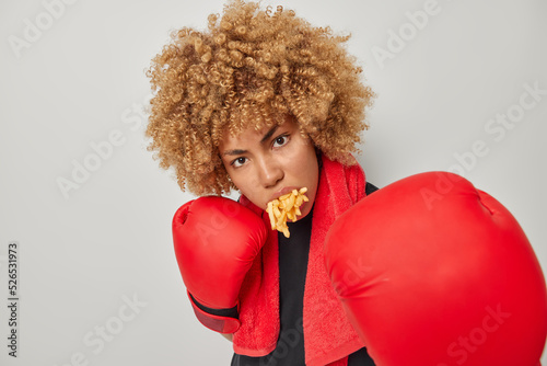 Fitness woman has mouth full of french fries stands in defensive pose exercises punches wears red boxing gloves clenches fists focused angrily at camera stands indoors. Boxing and sport concept photo