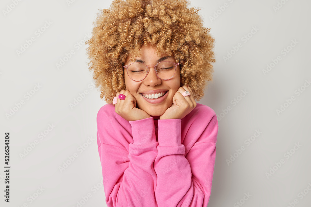 Indoor shot of pleased young woman with curly hair keeps eyes closed hands under chin smiles joyfully recalls pleasant memories wears spectacles and pink pullover isolated over grey background Indoor shot of pleased young woman with curly hair keeps eyes closed hands under chin smiles joyfully recalls pleasant memories wears spectacles and pink pullover isolated over grey background