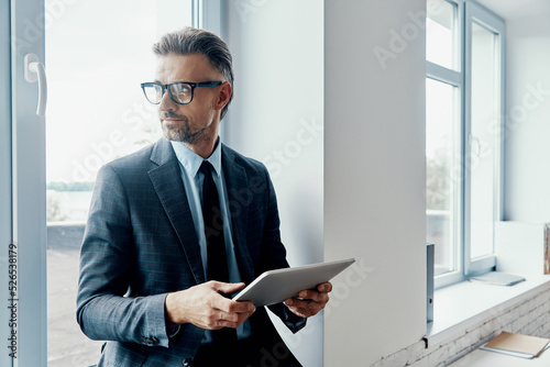 Thoughtful mature man in formalwear holding digital tablet while standing near the window in office