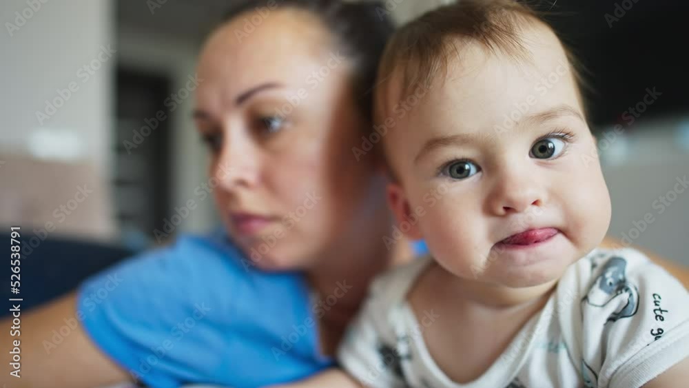 Mother and her baby boy beside. Little cite kid looks into camera with interest showing his tongue. Close up. Blurred backdrop. Mother and her baby boy beside. Little cite kid looks into camera with interest showing his tongue. Close up. Blurred backdrop.