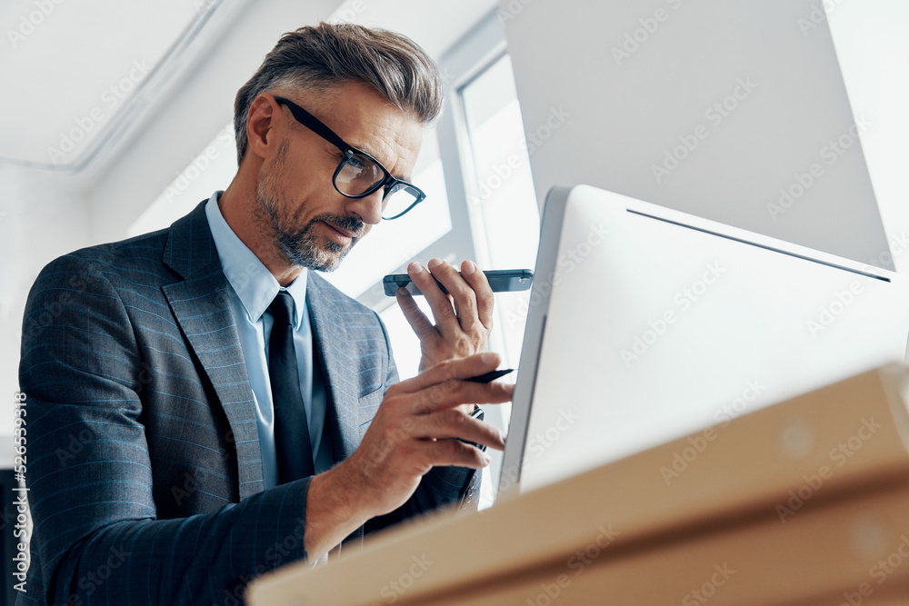 Concentrated businessman using loud speaker on phone while pointing computer monitor in the office Concentrated businessman using loud speaker on phone while pointing computer monitor in the office