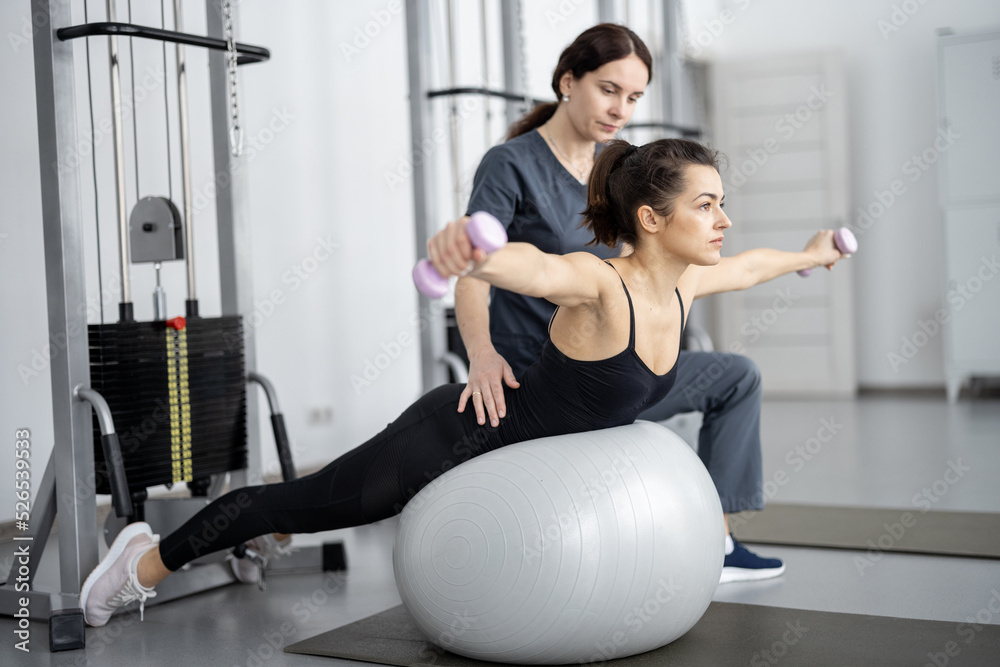 Obraz premium Woman doing exercises on fitness ball and dumbbells with rehabilitation specialist at the gym. Concept of physical therapy for back strengthening and recovery. Idea of recovery after pregnancy