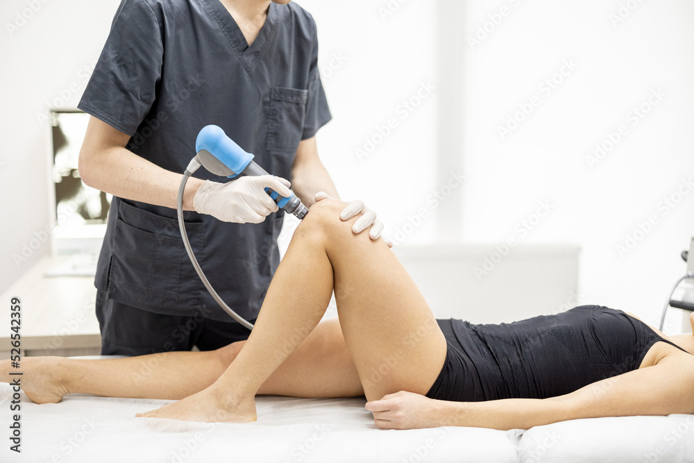 Doctor applies shock wave therapy with special medical equipment on women's knee joints at medical office. Concept of non-invasive technology for treating pain in musculoskeletal system