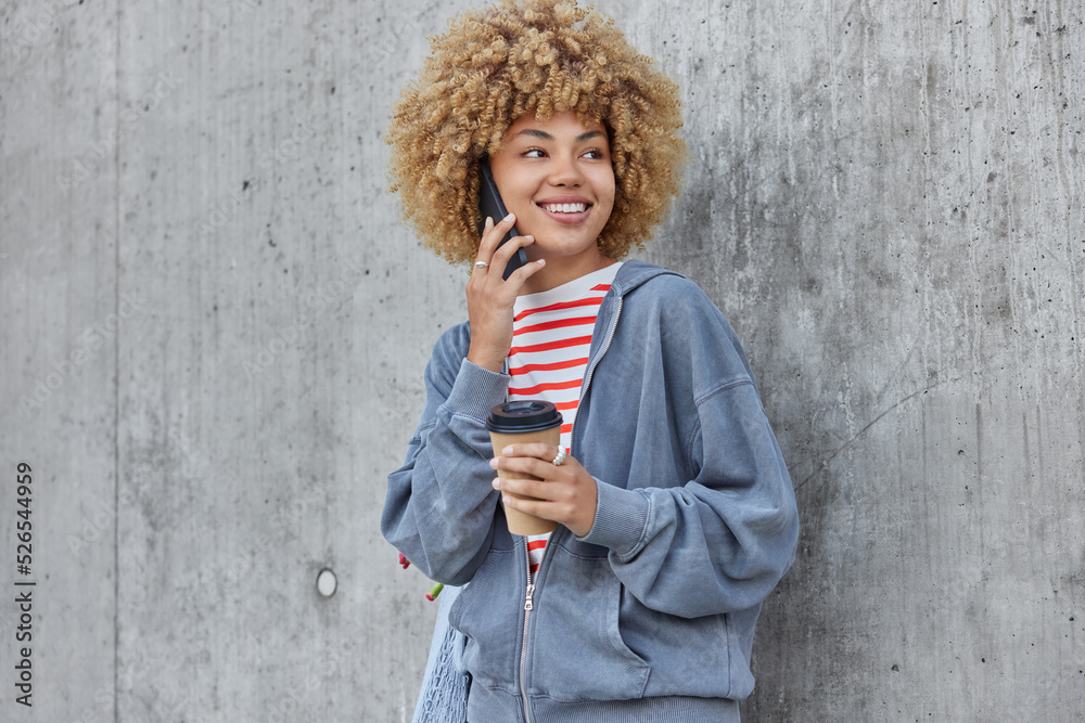 Fototapeta premium Young glad woman with curly hair makes phone call enjoys pleasant mobile conversation holds takeaway coffee focused aside dressed in casual hoodie poses over grey concrete wall. Distant communication