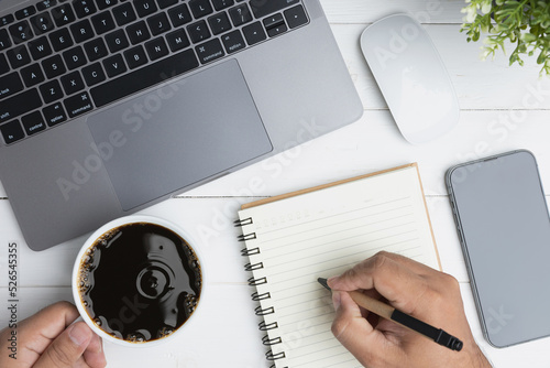 A hand man holding a white cup of coffee and writing on lank notebook. Top view the white desk office with a laptop, a mouse, and a coffee cup on a wood table. Flat lay top view copy space. photo