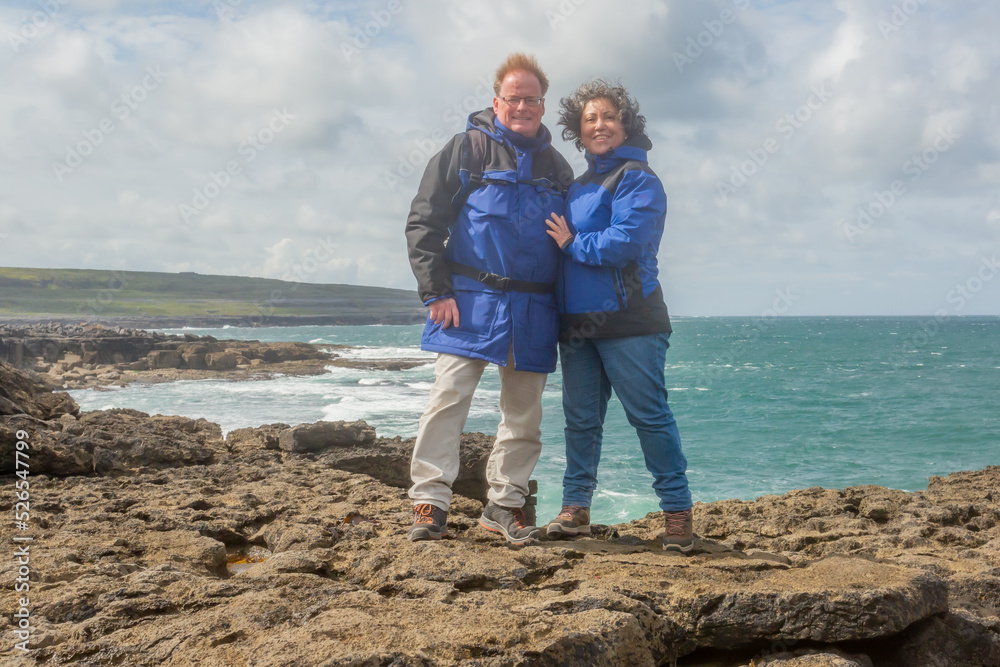 Senior adult couple standing on edge of the rocky cliff against the Atlantic ocean, blue sky with white clouds in blurred background, enjoying a spring day, blue jackets, island of Inis Oirr, Ireland