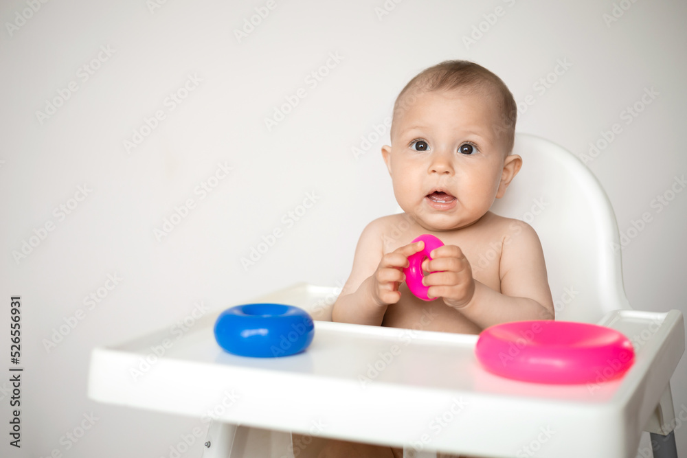 A cute little baby sits in a high chair and plays educational games. A light background, a place for text. A cute little baby sits in a high chair and plays educational games. A light background, a place for text.