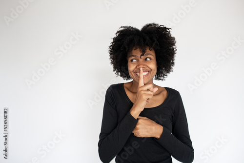 Portrait of cheerful African American woman with finger on lips. Young woman wearing black dress looking away and making silence gesture. Silence and secrecy concept