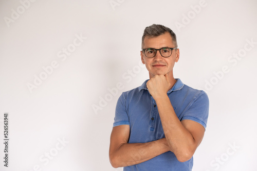 Portrait of confident mature man wearing glasses with hand on chin. Caucasian man wearing blue T-shirt looking at camera and smiling over white background. Success and eyesight concept