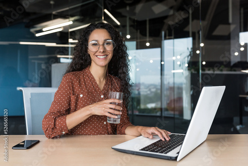 Portrait of happy and successful business woman inside modern office building, female worker smiling and looking at camera, holding glass of clean water, working on laptop at desk