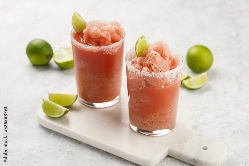 Two glasses with tall long watermelon drink with crushed ice, sugar rims, lime slices on marble board on grey background Two glasses with tall long watermelon drink with crushed ice, sugar rims, lime slices on marble board on grey background