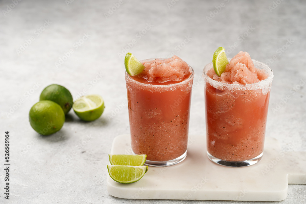 Two glasses with tall long watermelon drink with crushed ice, sugar rims, lime slices on marble board on grey background Two glasses with tall long watermelon drink with crushed ice, sugar rims, lime slices on marble board on grey background