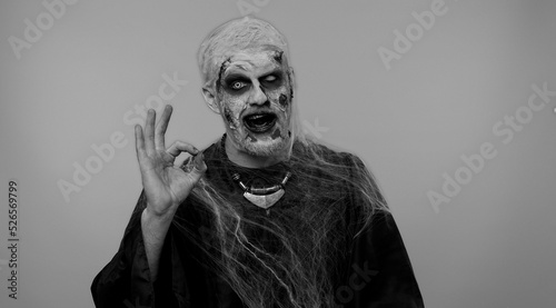 Creepy man with bloody scars face, Halloween stylish zombie make-up. Scary wounded undead guy looking approvingly at camera showing Ok gesture, like sign positive something good and smiles terribly
