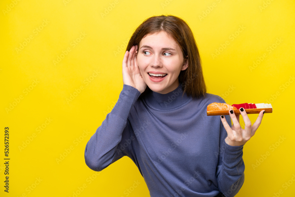 Young English woman holding sashimi isolated on yellow background listening to something by putting hand on the ear Young English woman holding sashimi isolated on yellow background listening to something by putting hand on the ear
