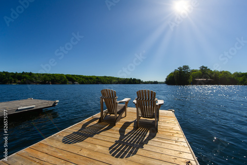 Cottage life - Sunrise on two empty Adirondack chairs sitting on a dock on a lake in Muskoka, Ontario Canada. The sun light create long shadows on the wooden pier.