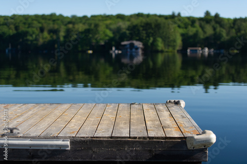 Empty portion of a wooden dock facing a lake in Muskoka Ontario, Canada. Across the the calm waters some cottages are visible nestled between green trees. 