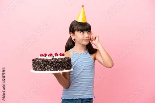 Little caucasian kid holding birthday cake isolated in pink background having doubts and thinking