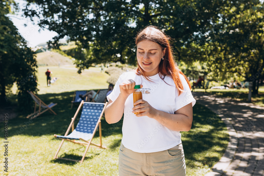 Fototapeta premium Beautiful woman in sunglasses drinking fresh juice outdoors. Redhead girl walking in the park and holding a big bottle of fresh cold smoothie in a warm sunny day. Multivitamin Juice Bottle Mockup