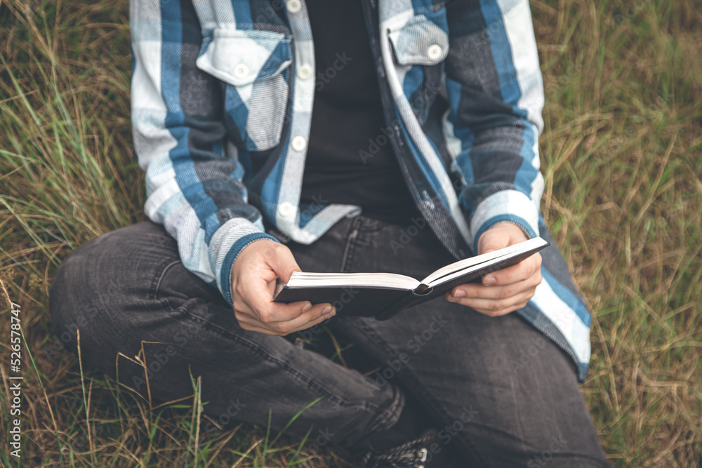 A man in a plaid shirt reads a book in nature.